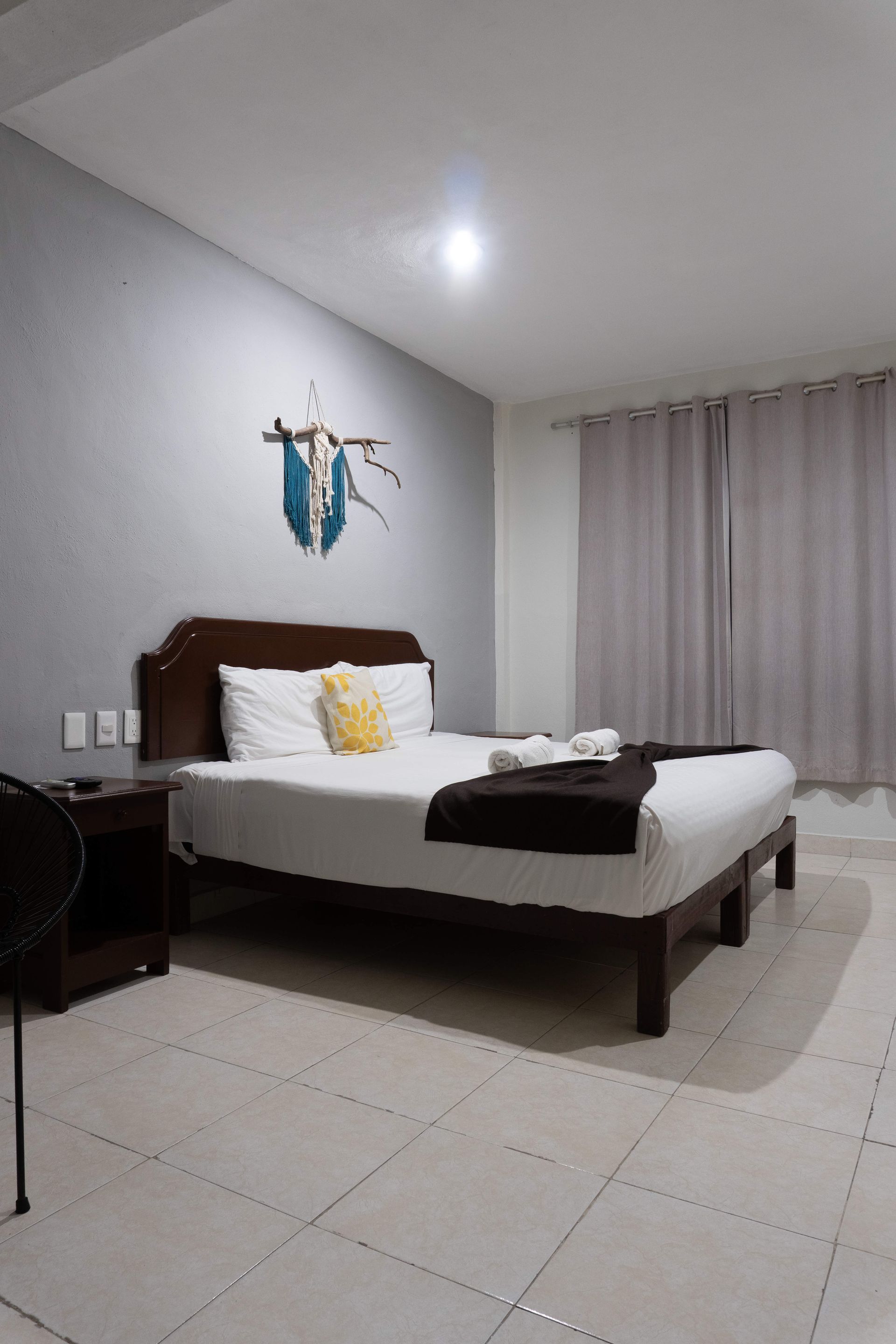 Habitación espaciosa con cama, suelo de baldosas blancas, cortinas grises y decoración en la pared sobre el cabecero.