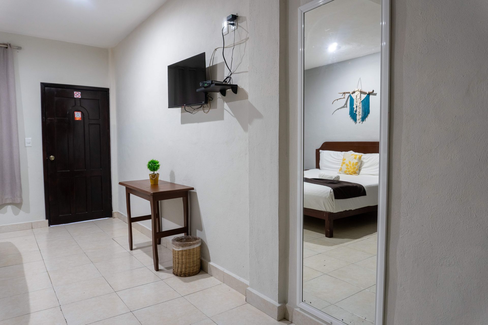 Pasillo luminoso con televisor montado en la pared, mesa pequeña, cesta y espejo que refleja una cama en un dormitorio.