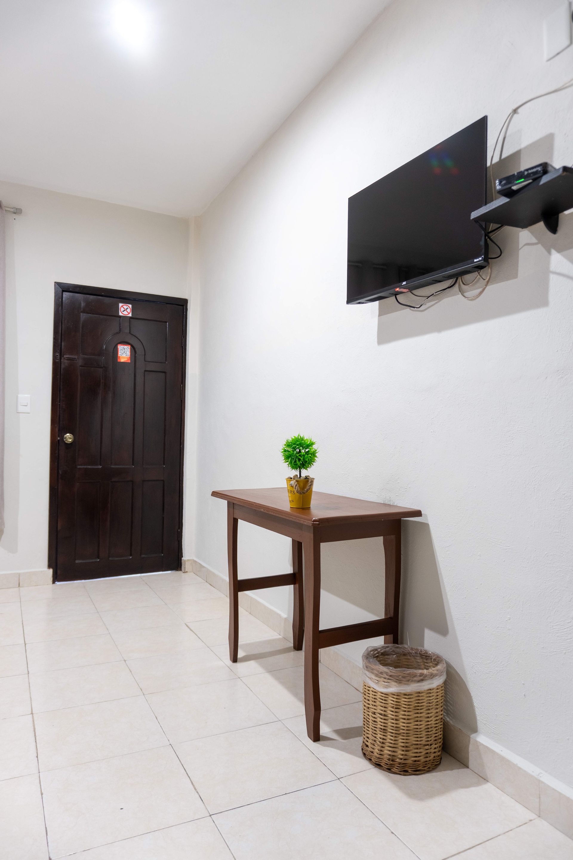 Pequeño pasillo blanco con puerta de madera oscura, TV en pared, mesa estrecha, planta decorativa y cesta de mimbre.
