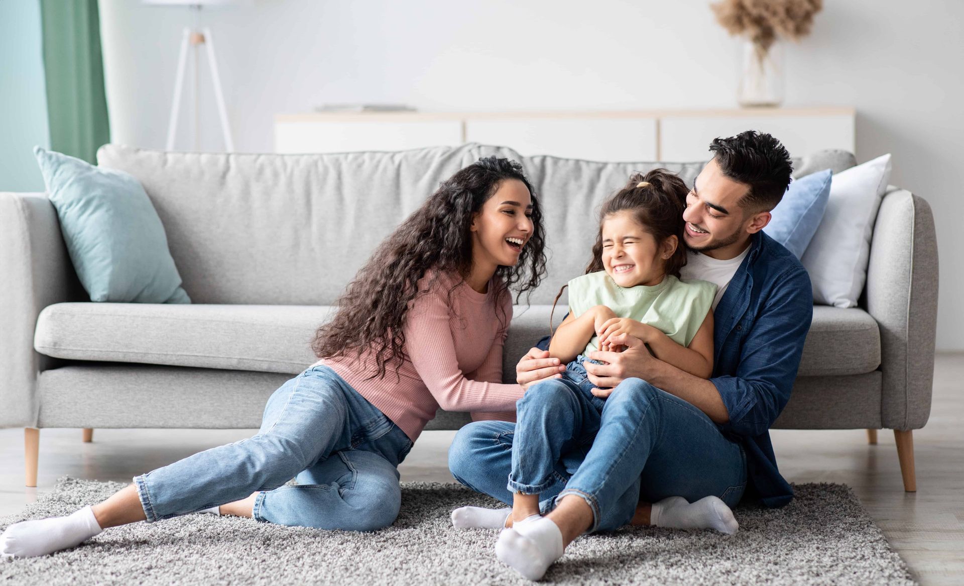 Family sitting on a living room rug, smiling and cuddling in front of a gray sofa.