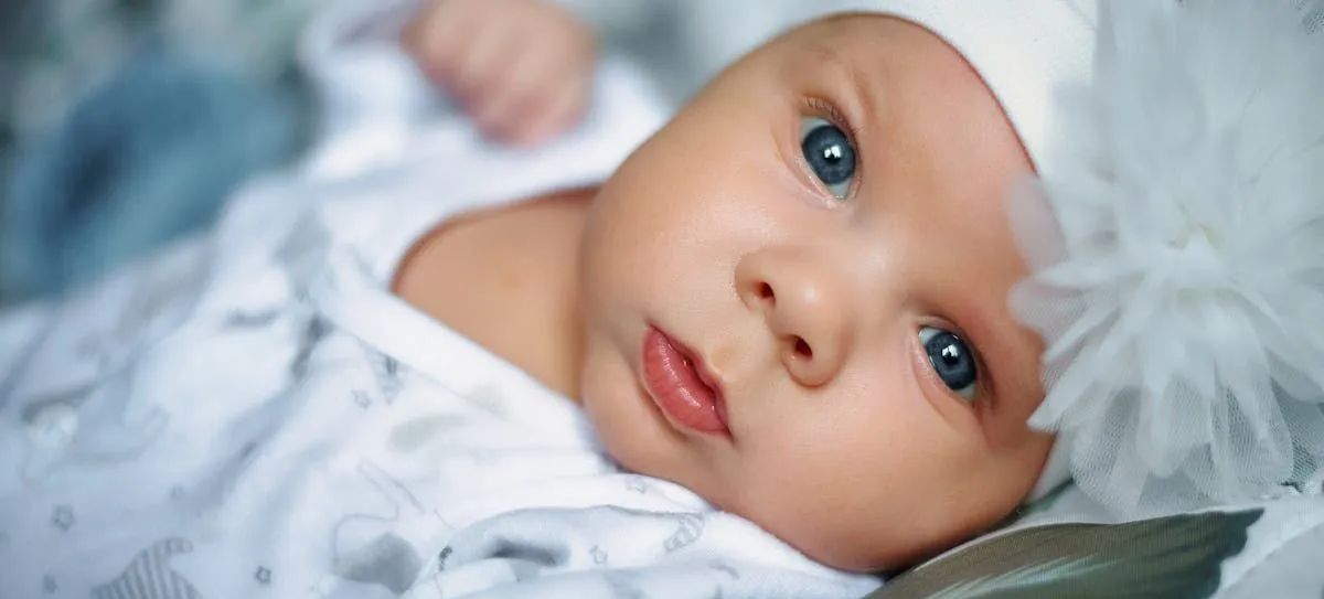 A baby is wearing a white hat and looking at the camera.