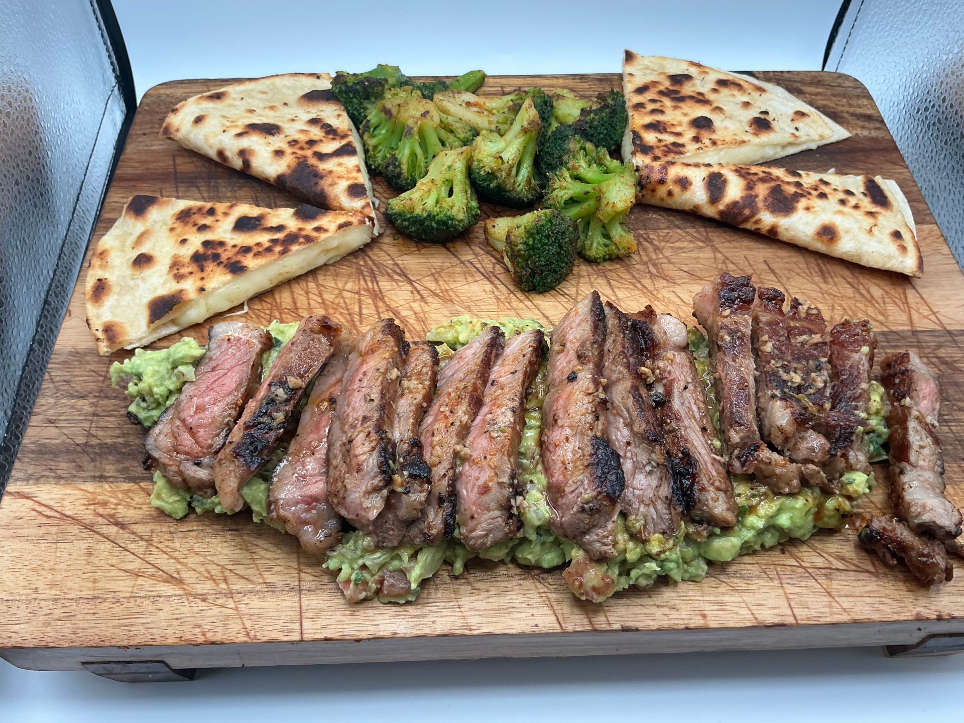 Grilled steak, broccoli, and naan bread arranged on avocado mash, on a wooden board.