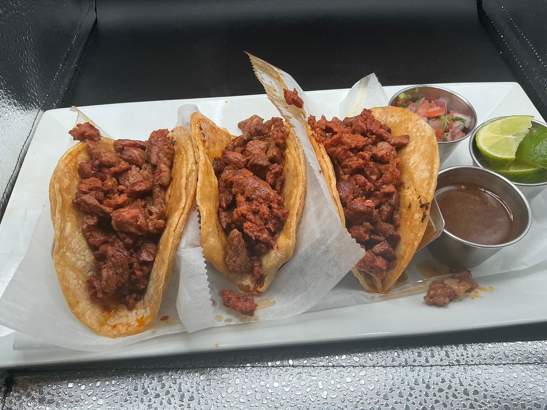 Three tacos filled with meat, served on a white plate with salsa, lime wedges, and dipping sauce.