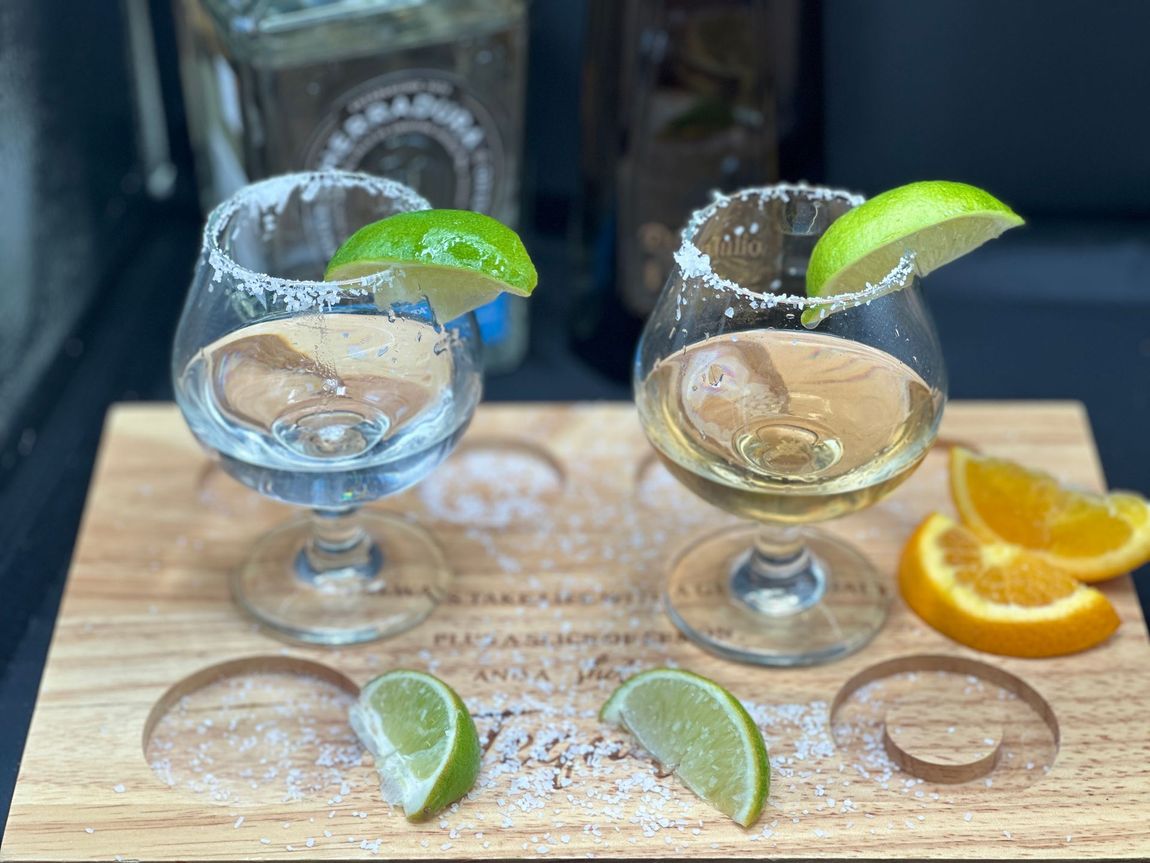 Two tequila shots with salt rims and lime wedges on a wooden board with citrus.