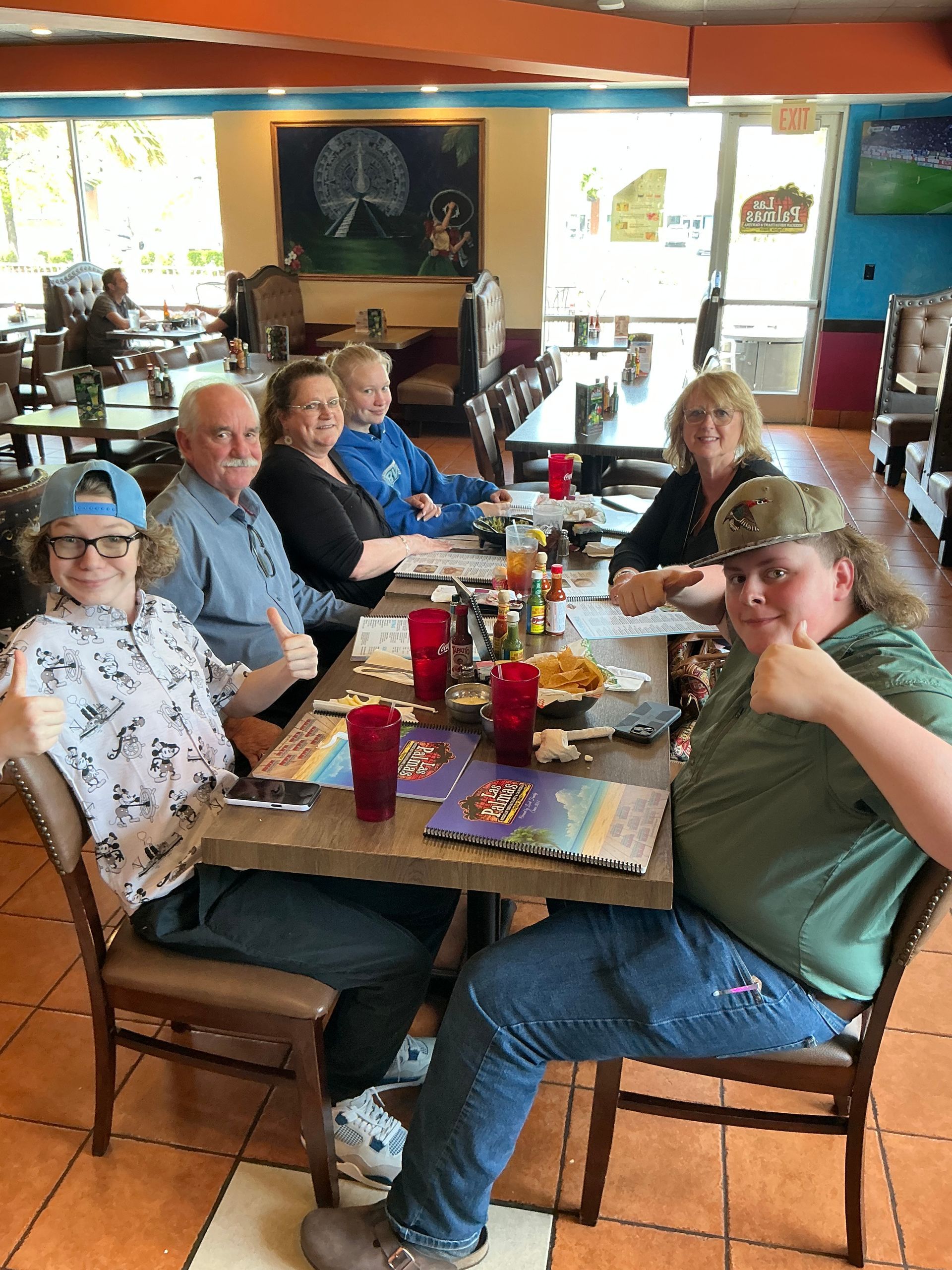 Six people at a restaurant table, smiling, giving thumbs-up. Mexican restaurant interior; colorful decor.