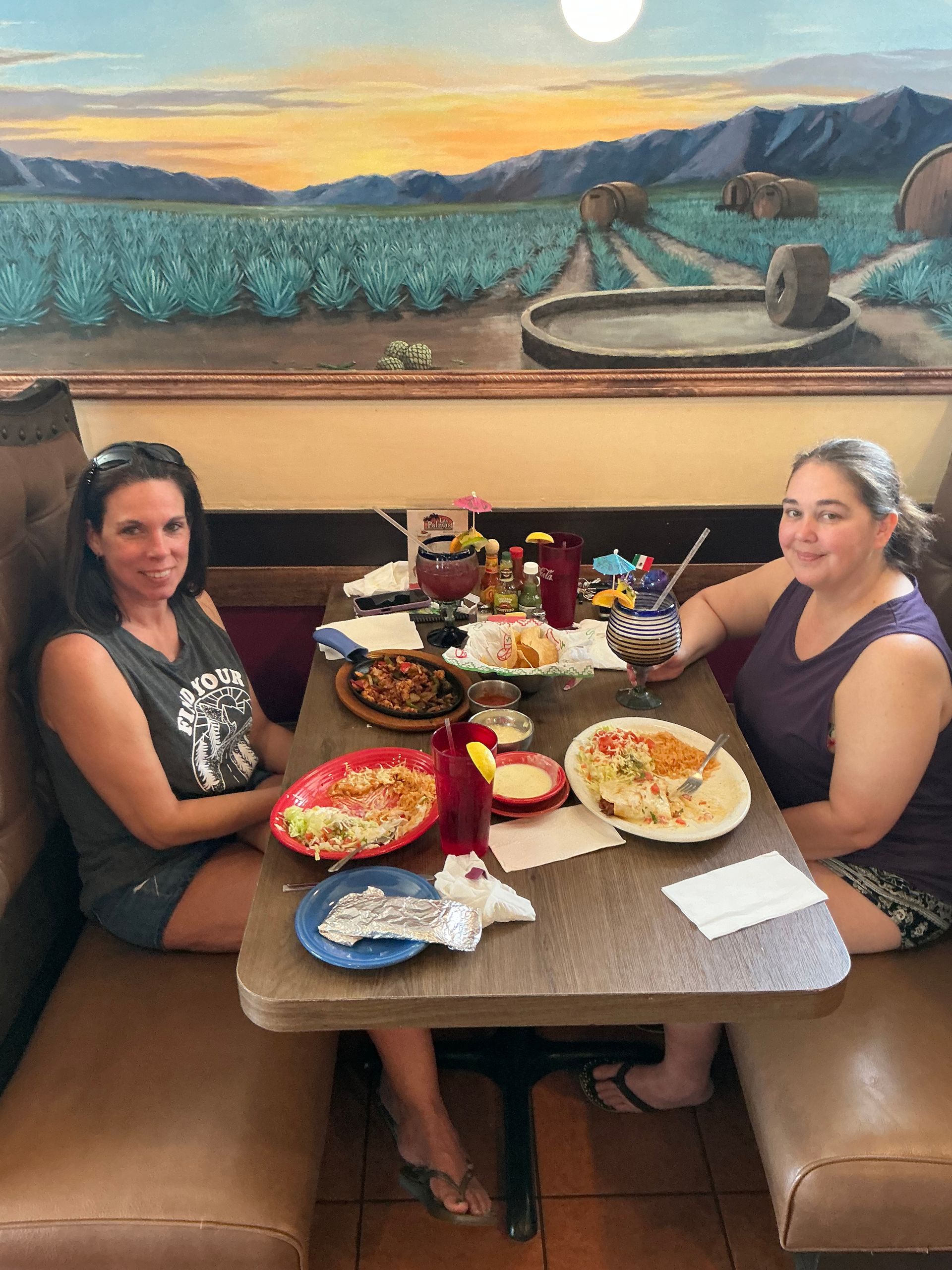Two people seated at a table in a restaurant with food and drinks. Background is a mural.