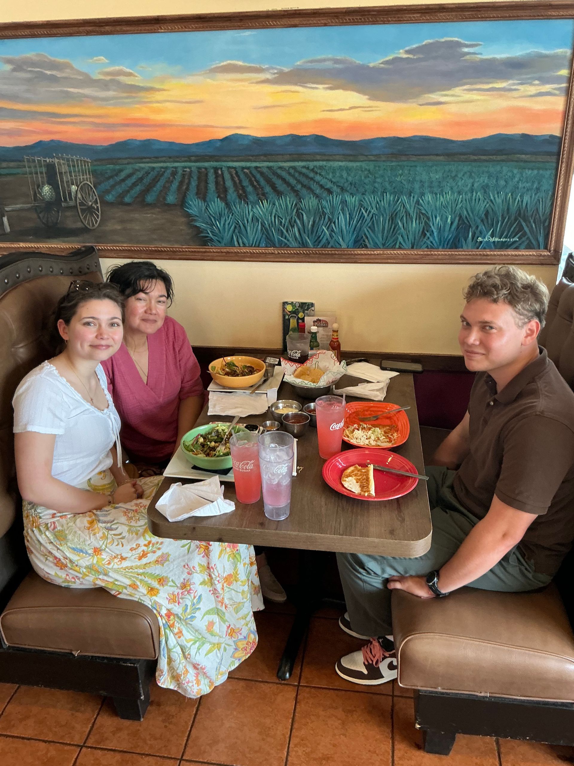 Three people seated at a restaurant table. One is in a white shirt, one in a pink shirt, and the last in a brown shirt. Food on table.