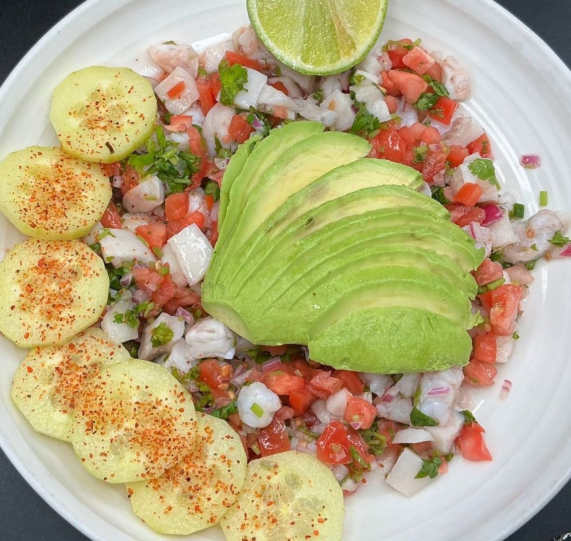 Ceviche with avocado, cucumber slices, and lime wedge on a white plate.