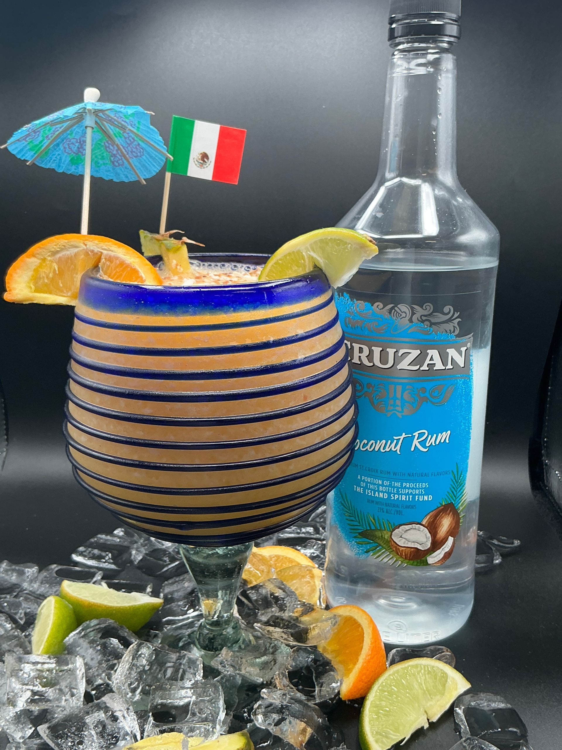 Cocktail in blue-striped glass, decorated with citrus, umbrella, Mexican flag, next to a rum bottle on ice.