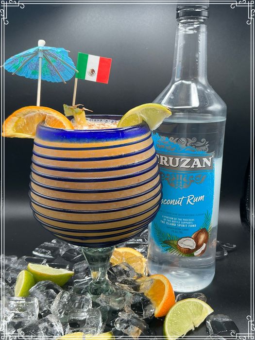 Cocktail in blue-striped glass, with coconut rum bottle, orange slices, and Mexican flag.