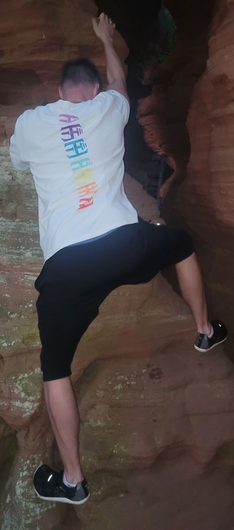 Een persoon beklimt een rode rotswand, gekleed in een wit T-shirt, zwarte korte broek en sportschoenen.