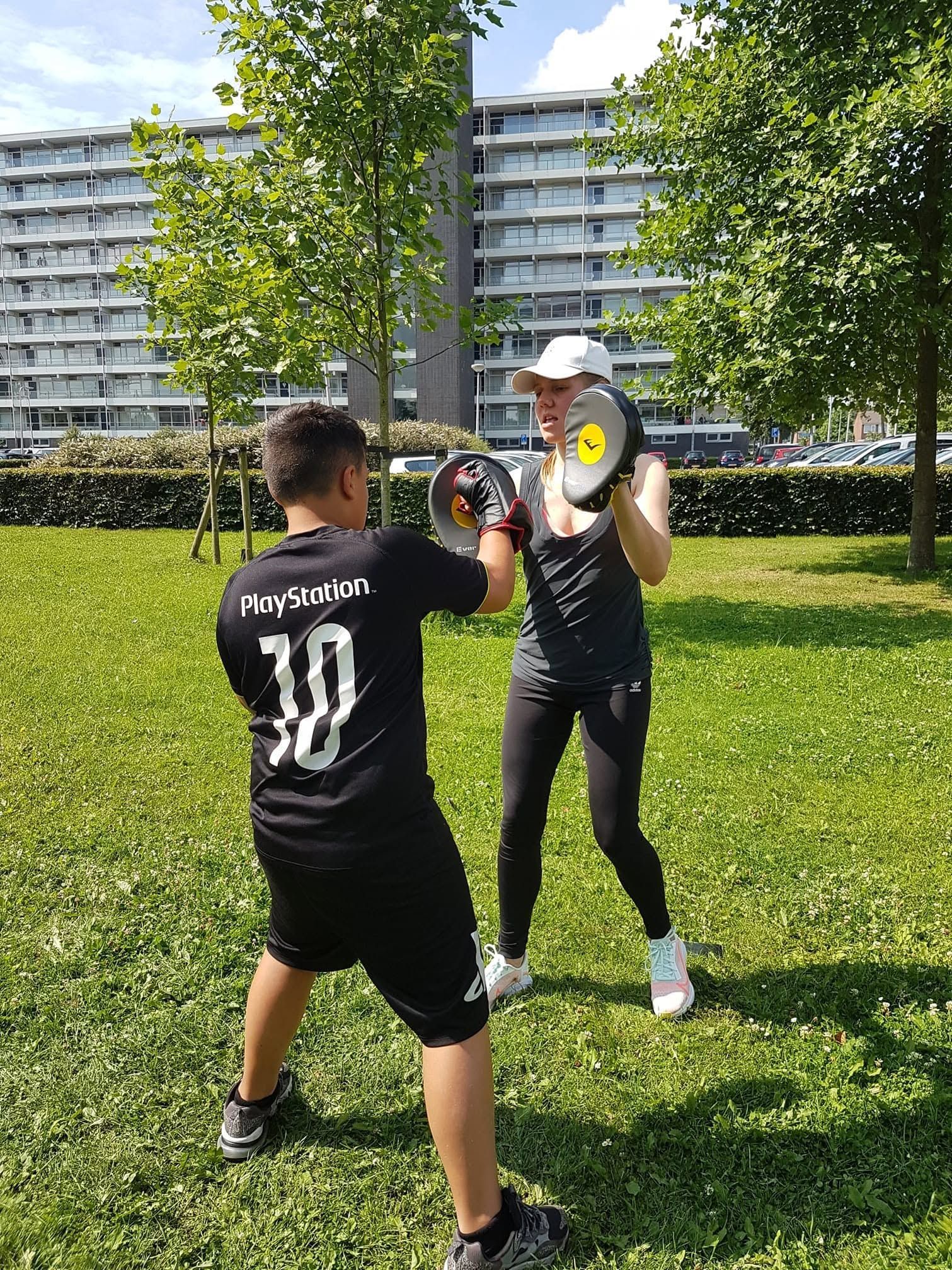 Twee mensen, gekleed in zwarte sportkleding, oefenen boksoefeningen op een grasveld in de buurt van appartementencomplexen.