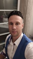 Selfie van een persoon in een wit shirt en een blauw vest, staand binnen bij een raam met gordijnen.