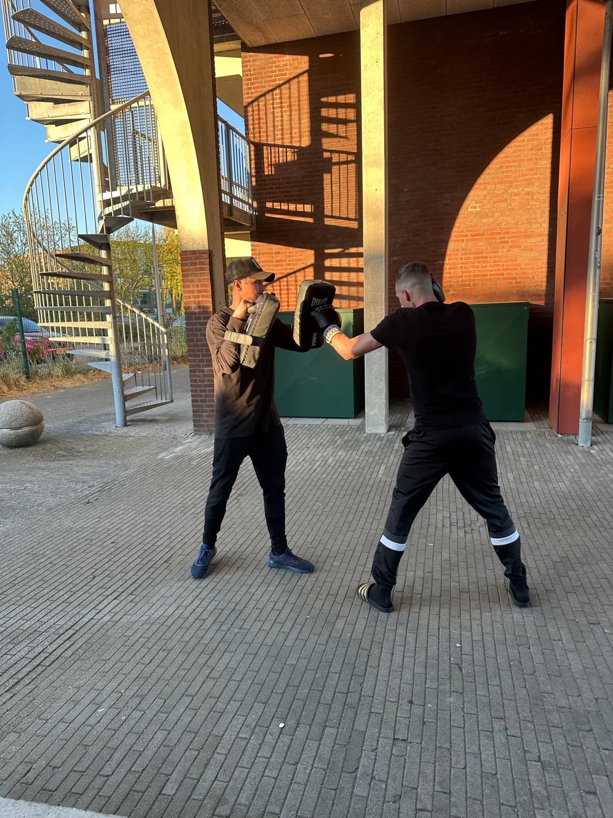 Twee mensen beoefenen vechtsporten in de buitenlucht, vlakbij een bakstenen gebouw en een metalen wenteltrap.