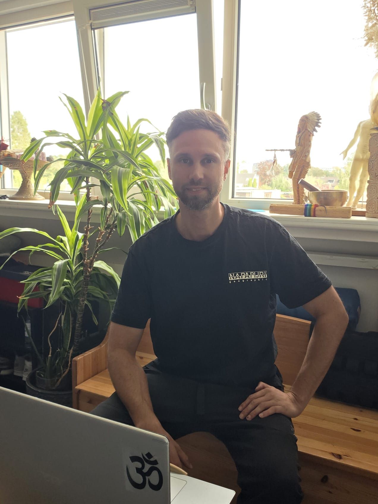 Een man zit binnen naast een laptop, met grote potplanten en een licht raam achter hem.