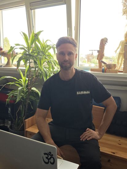 Een man zit binnen naast een laptop, met grote potplanten en een licht raam achter hem.