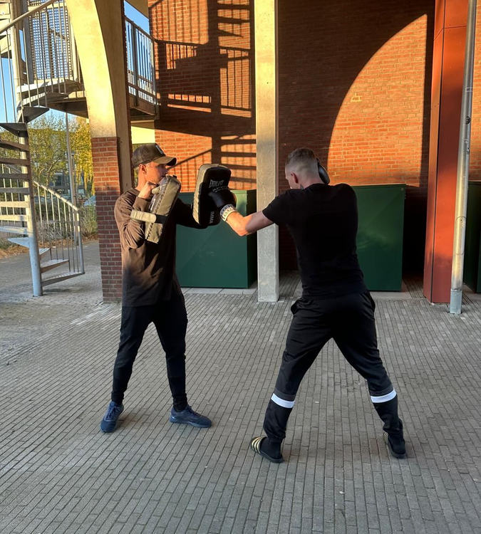 Twee mensen sparren met bokshandschoenen onder een bakstenen boog in de buitenlucht.