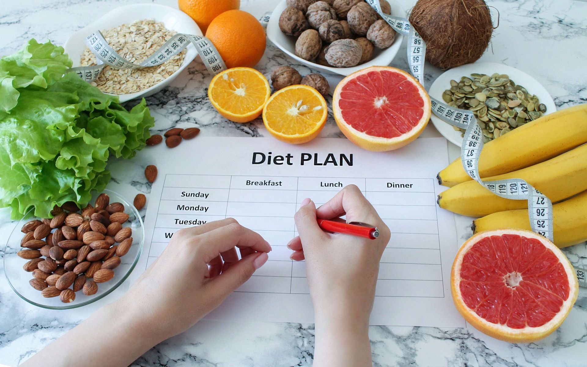 Person writing on a diet plan surrounded by healthy foods: oranges, grapefruit, nuts, bananas, seeds, and lettuce.