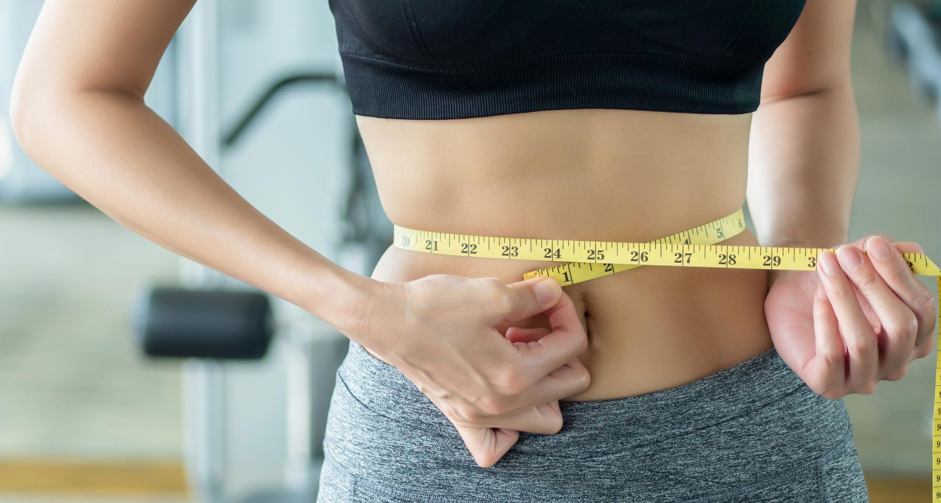Woman in sports bra and leggings measuring her waist with a yellow tape measure in a gym.
