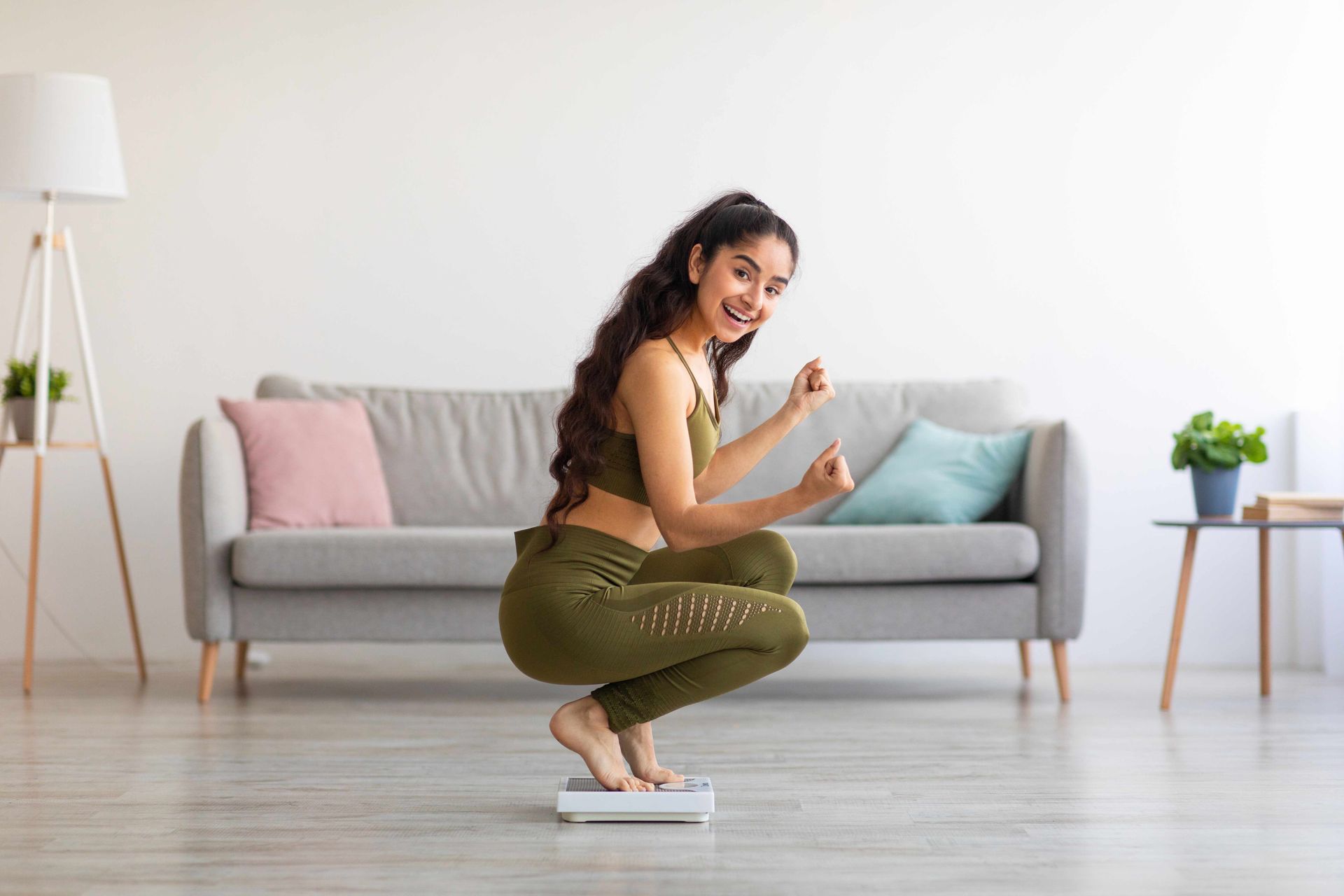 Woman in olive green workout clothes squats on a scale, arms raised in excitement. Living room setting.