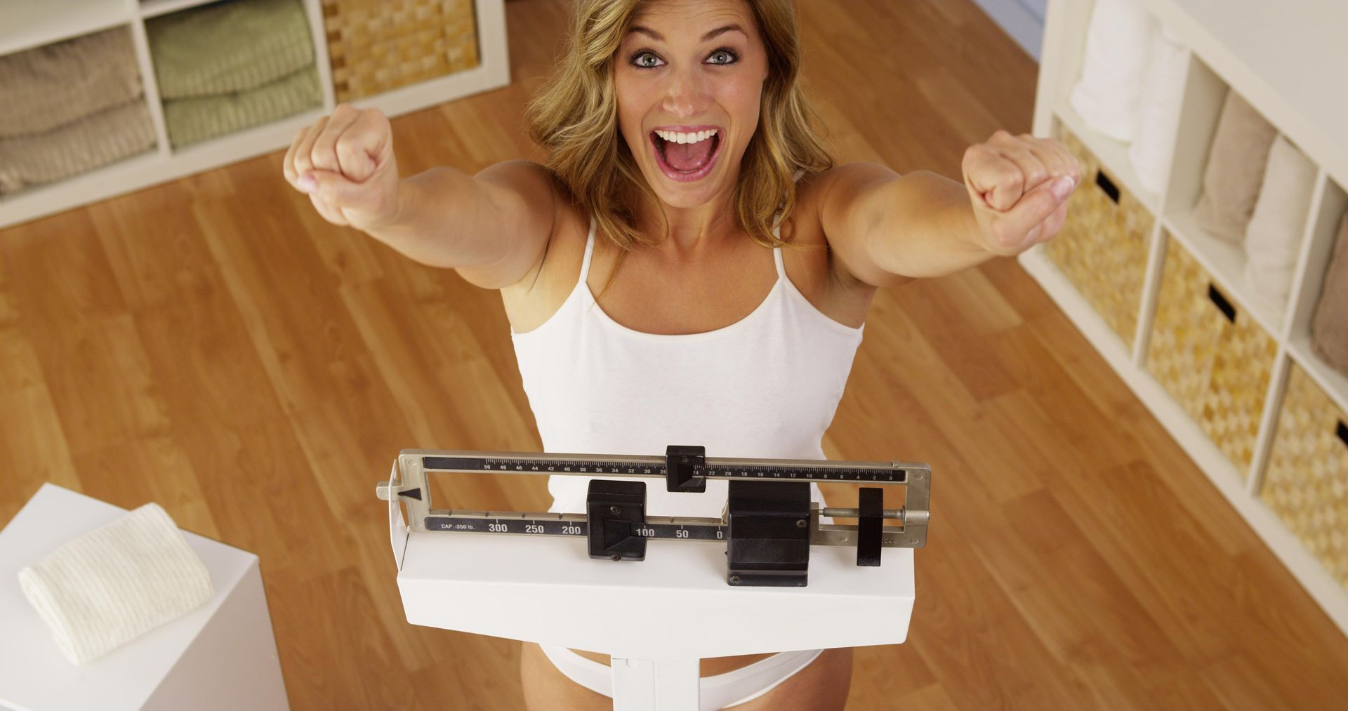 Woman cheers with arms raised while standing on a scale in a brightly lit room.