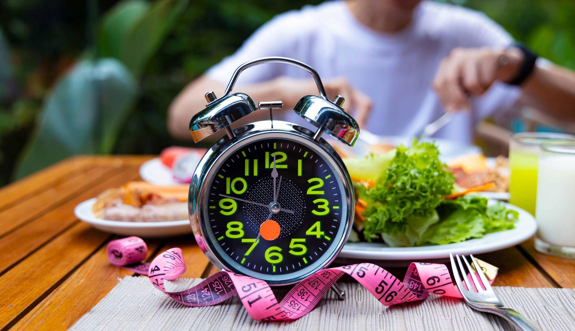 Alarm clock with food plates and measuring tape. Person eating in the background.