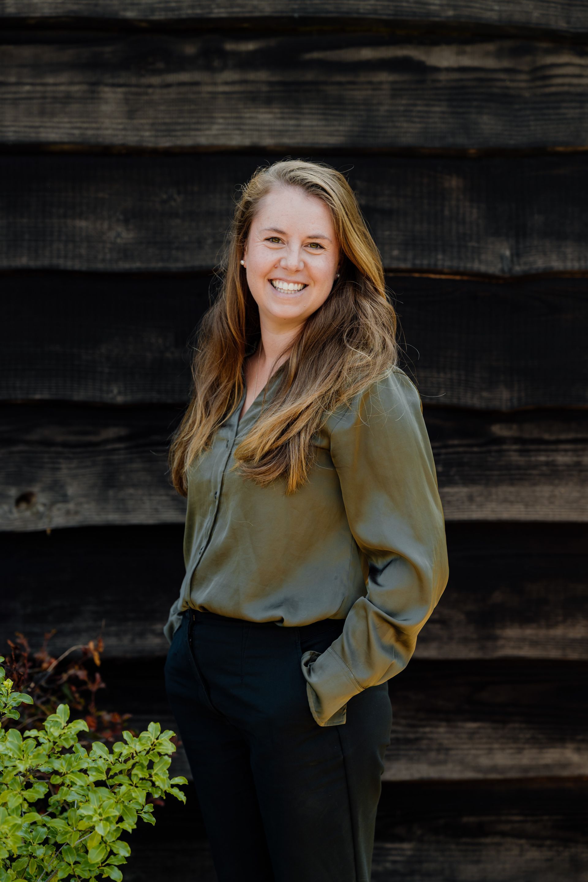 A woman in a green shirt and black pants is standing in front of a wooden wall.