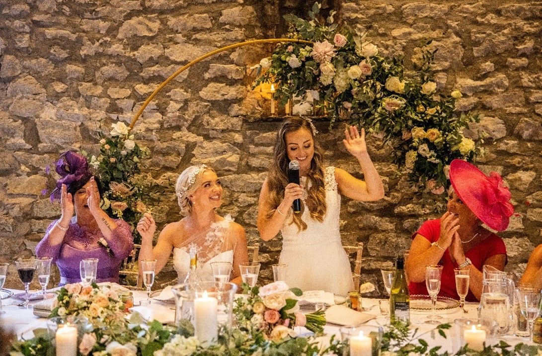 A bride is giving a speech to her wedding guests while sitting at a table.