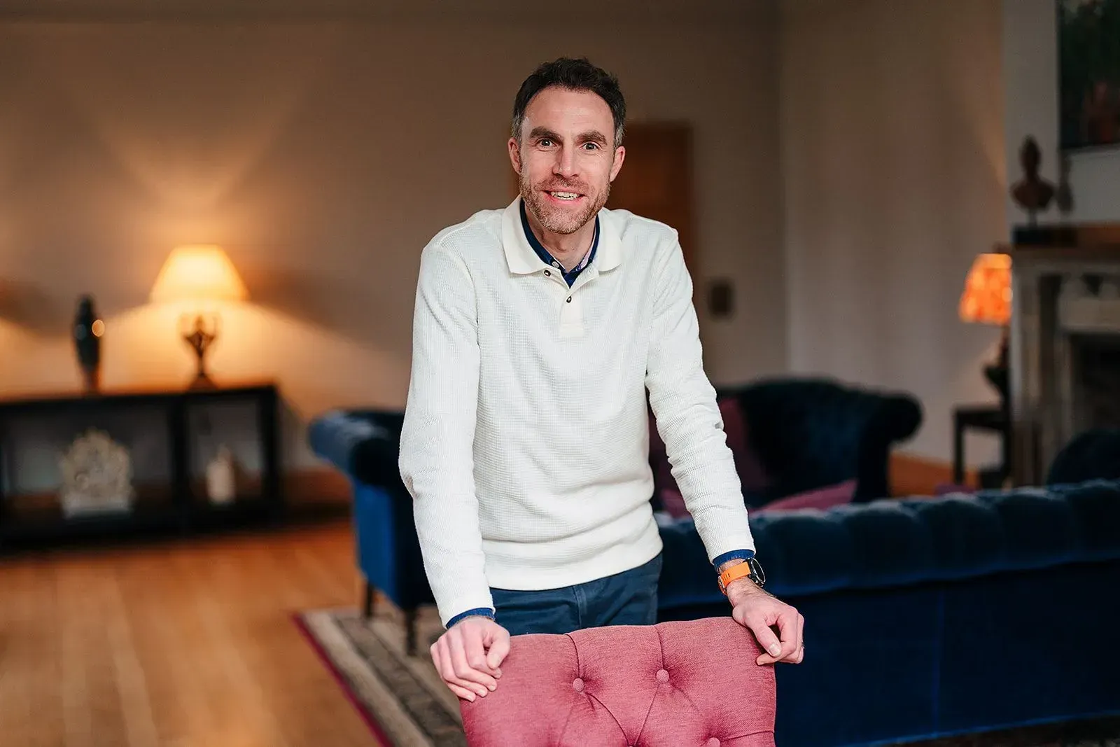 A man in a white sweater is standing next to a pink chair in a living room.