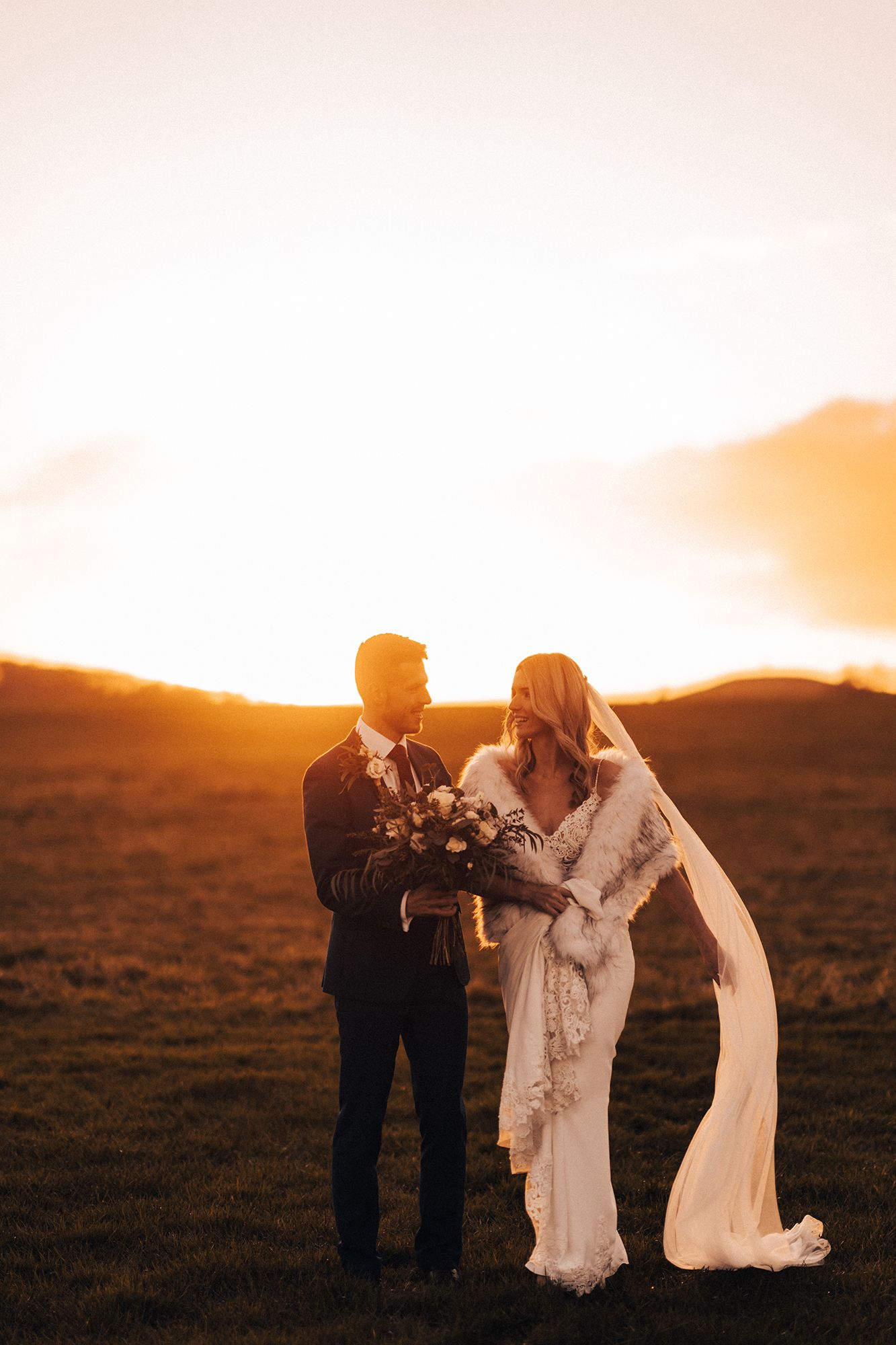A bride and groom are standing in a field at sunset.