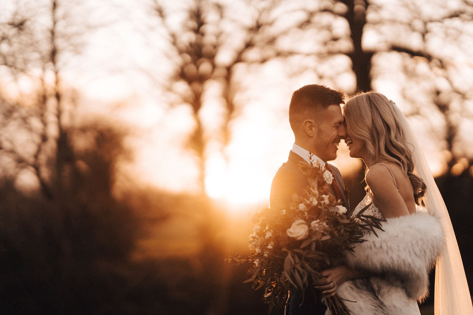 A bride and groom are standing next to each other in a field at sunset.