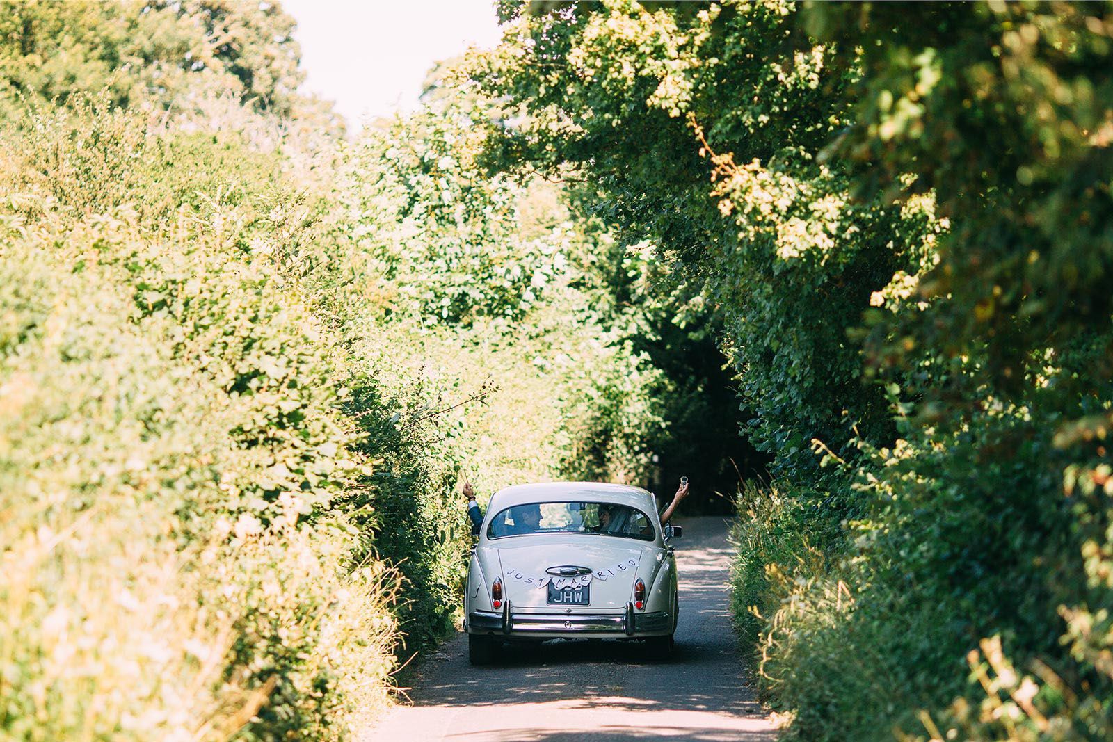 A white car is driving down a country road surrounded by trees.