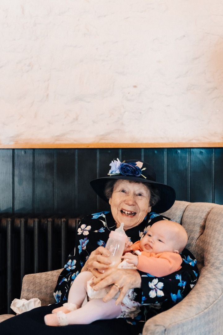 An elderly woman is sitting in a chair holding a baby and a bottle.