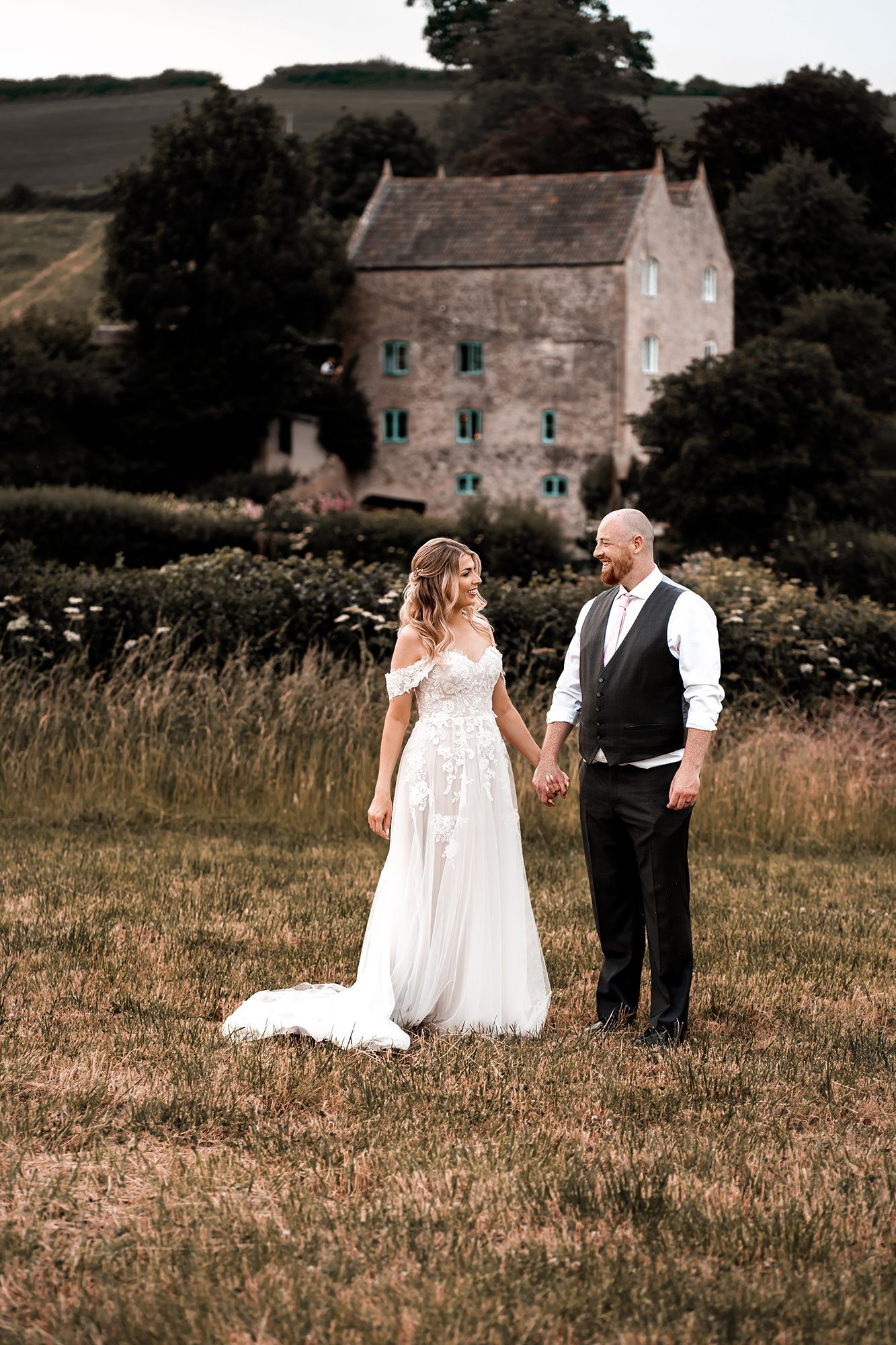 A bride and groom are holding hands in a field in front of a building.