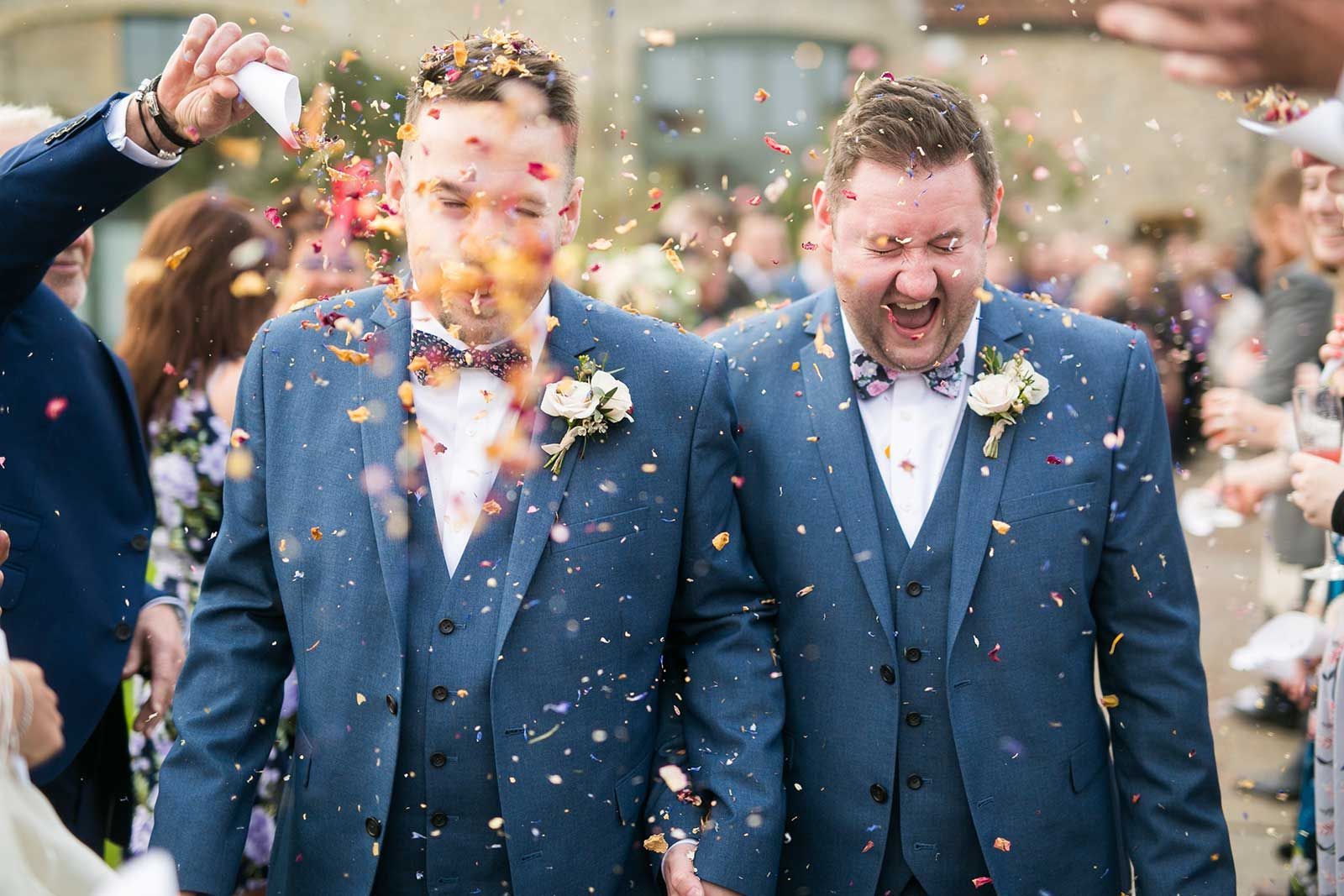 Two men in suits are walking through confetti at a wedding.