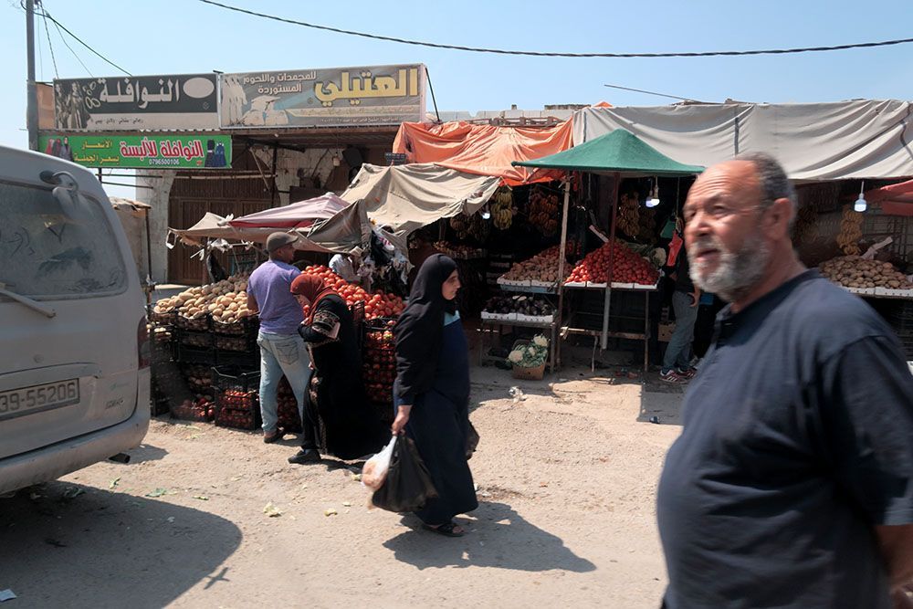 Street market with produce stalls, people shopping. Man in blue shirt, woman in black dress, van.