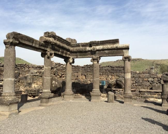 Ruins of an ancient synagogue with dark stone columns and lintels. Brown stones in the background, set against a blue sky.
