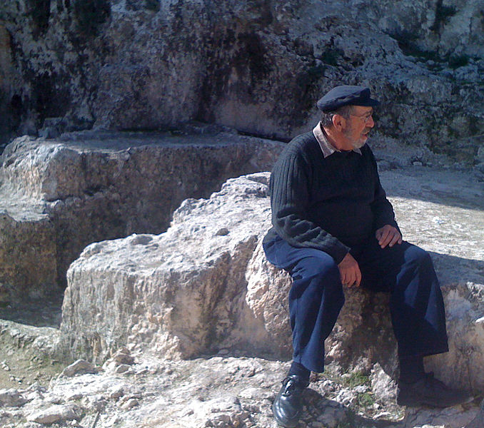 Man in dark sweater and hat sits on large rocks outdoors, looking to the side.