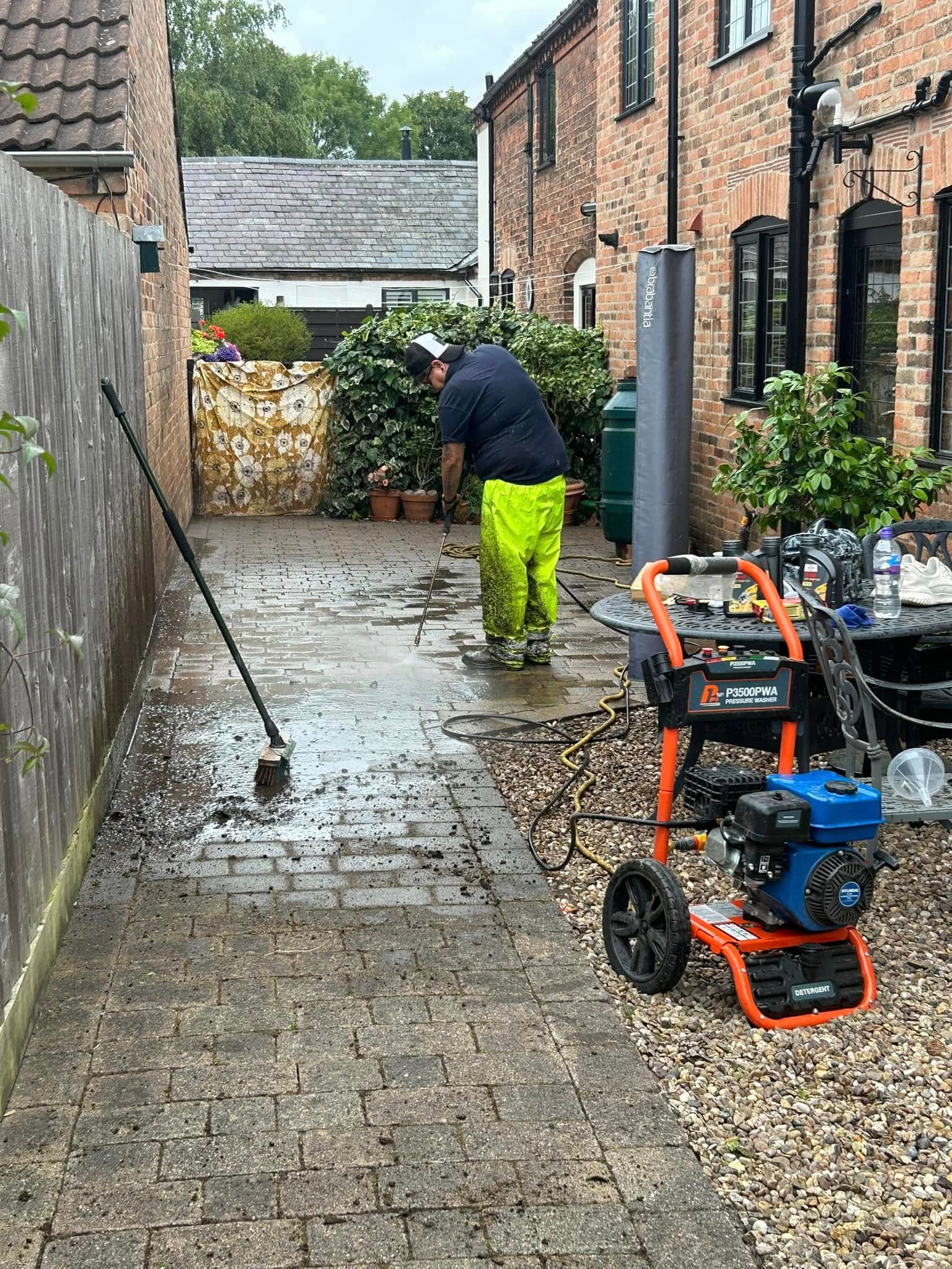 Person pressure-washing a wet driveway beside brick buildings, with equipment in the foreground.