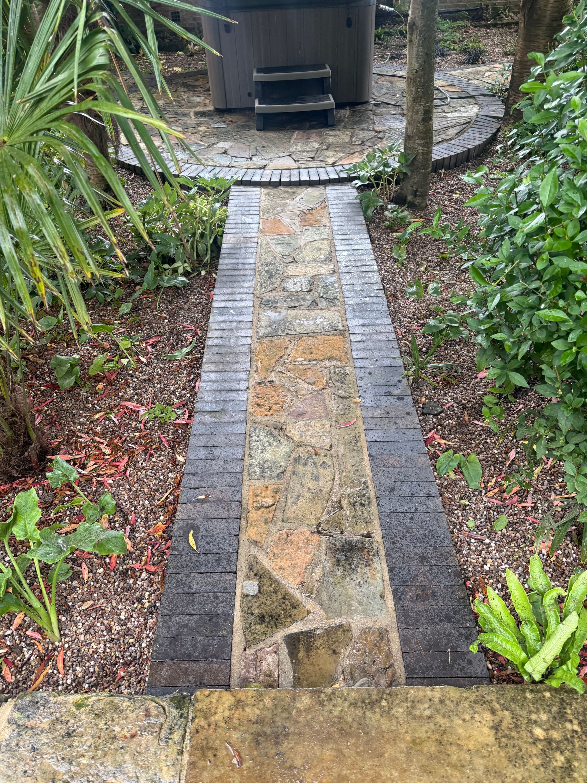 Narrow stone garden path leading to a small wooden bench in a lush, wet landscape.