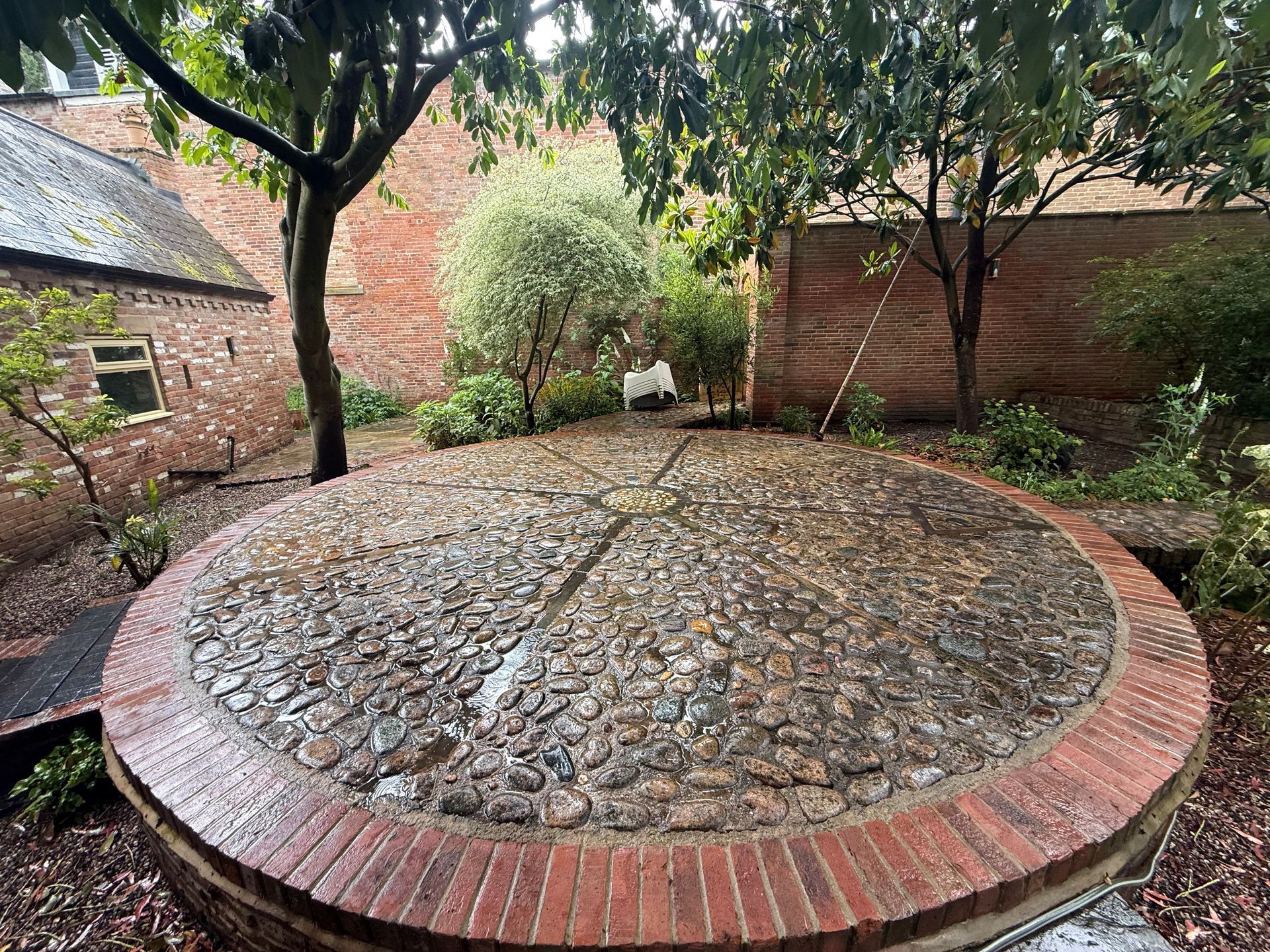 Circular cobblestone patio bordered by red bricks in a leafy garden courtyard.