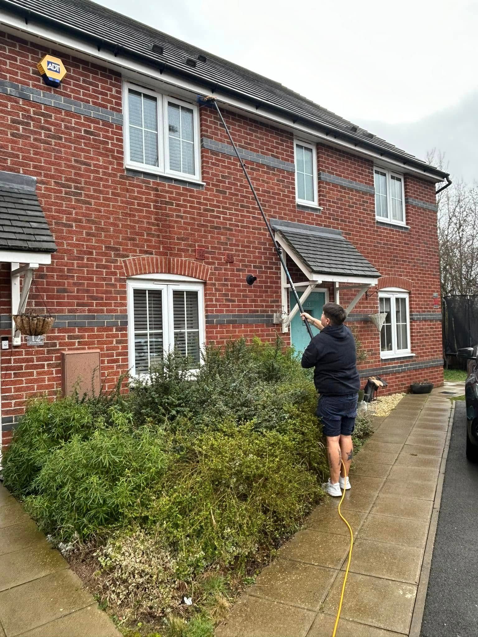 Person on a wet sidewalk washing a red brick house with a hose