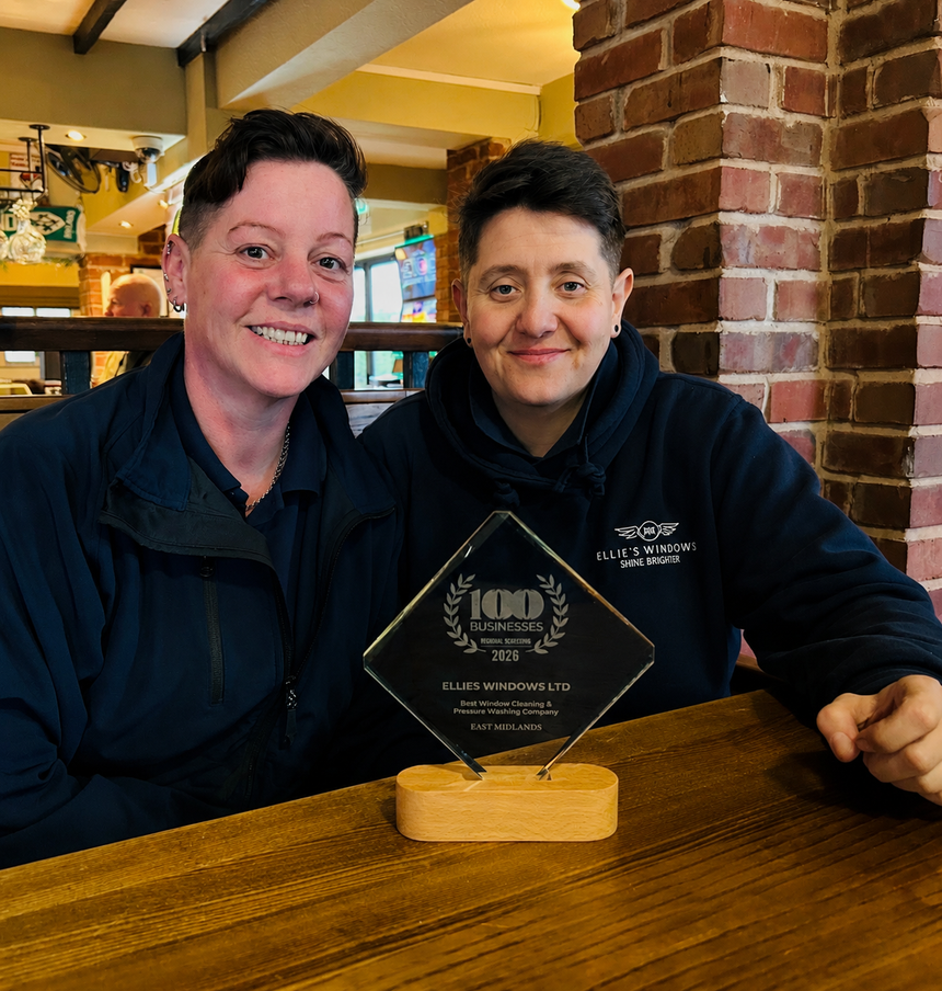 Two people smiling behind a glass award plaque at a wooden table in a brick-walled restaurant.