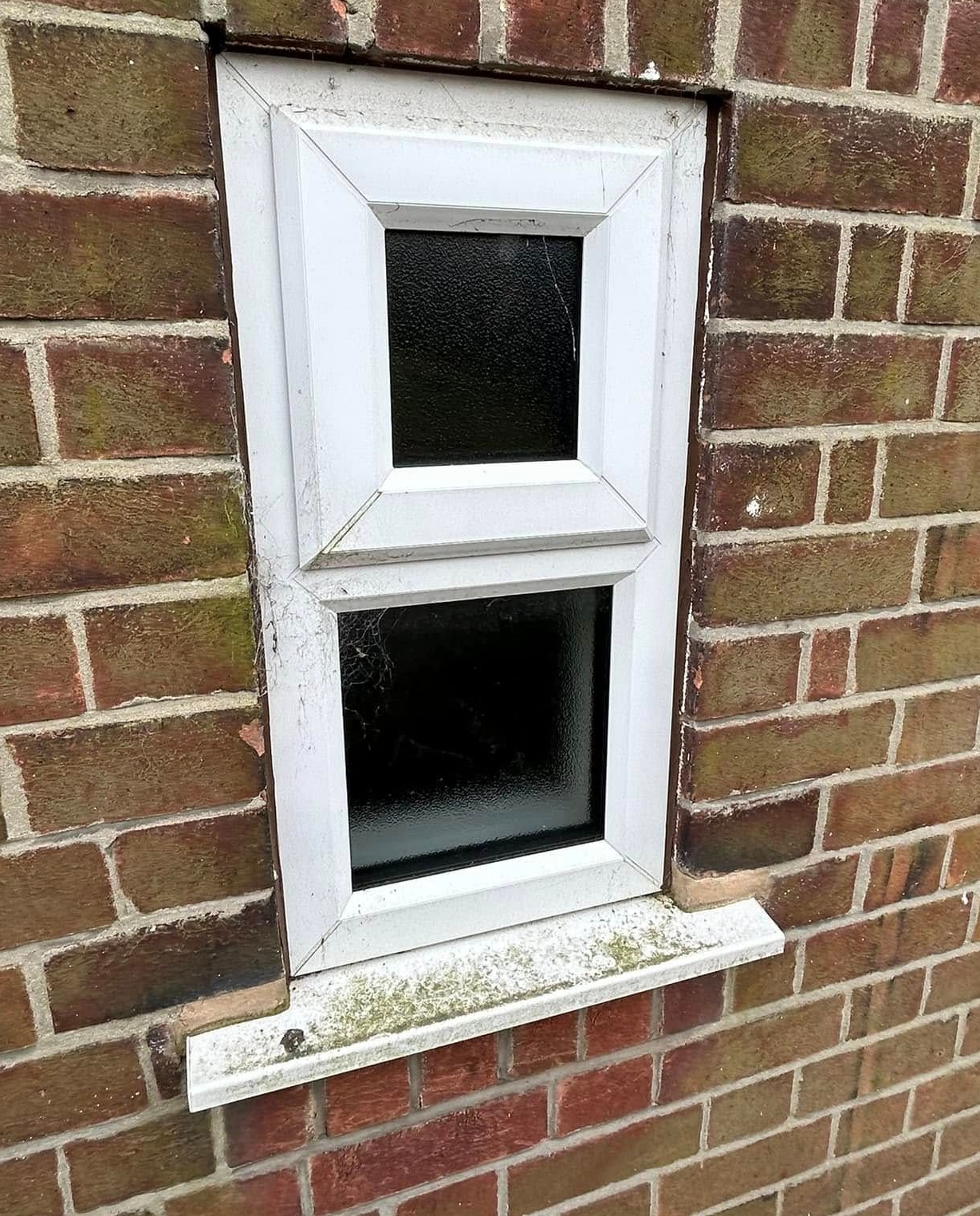 Small white-framed window set in a red brick wall, with a small lower ledge and dark glass panes.