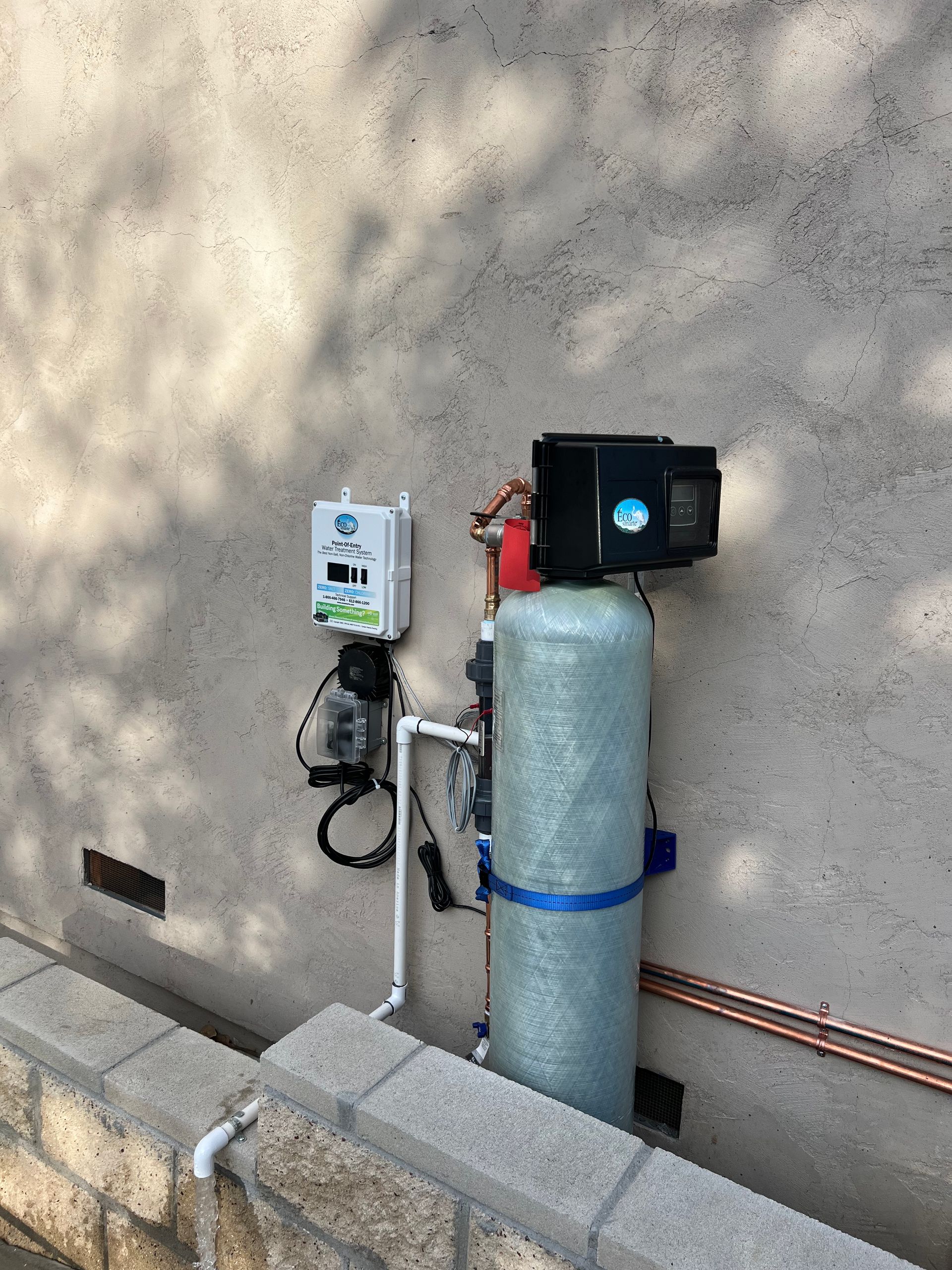 Water filtration system mounted on a stucco wall outside.  Blue tank, black box, and white control panel with plumbing.