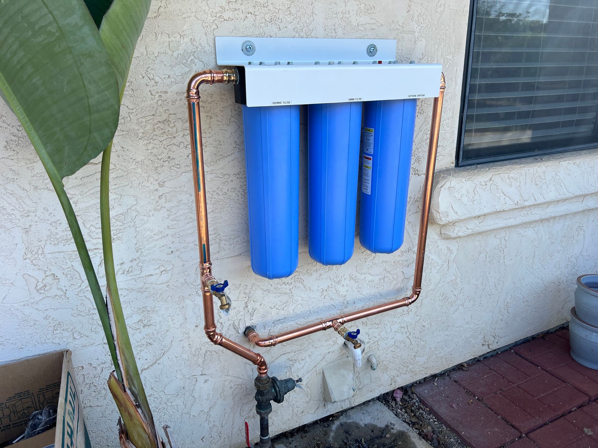 A triple-filter water system with copper pipes mounted on an exterior wall.