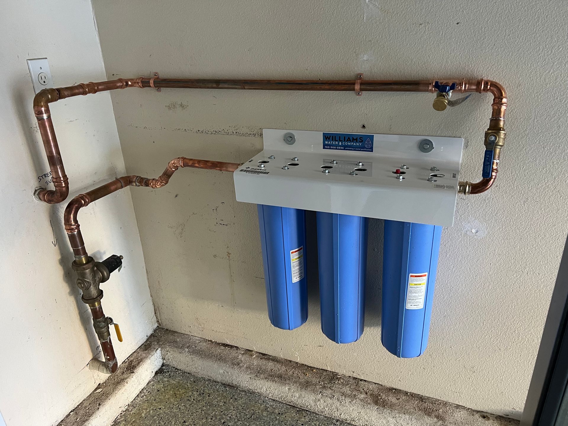Copper pipes connected to a three-stage water filter system on a beige wall in a garage.