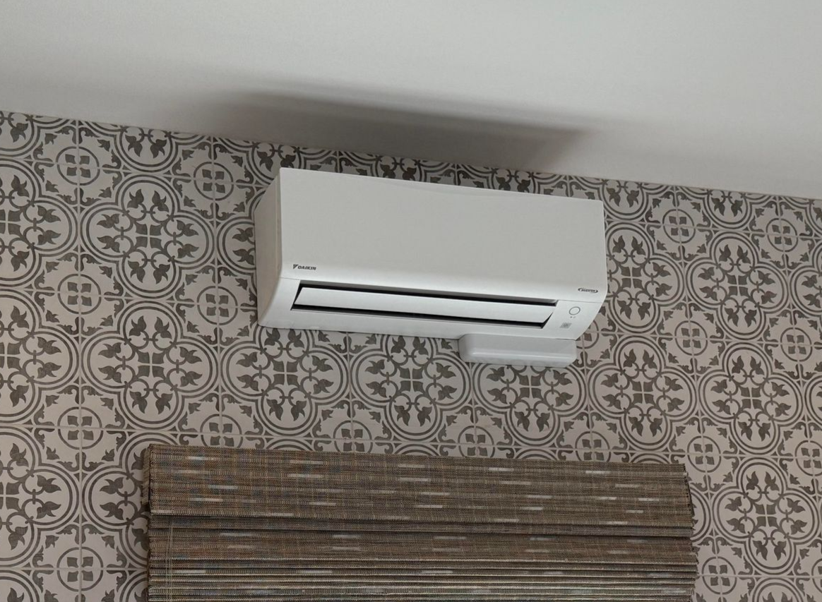 White wall-mounted air conditioner blowing cool air over a window.
