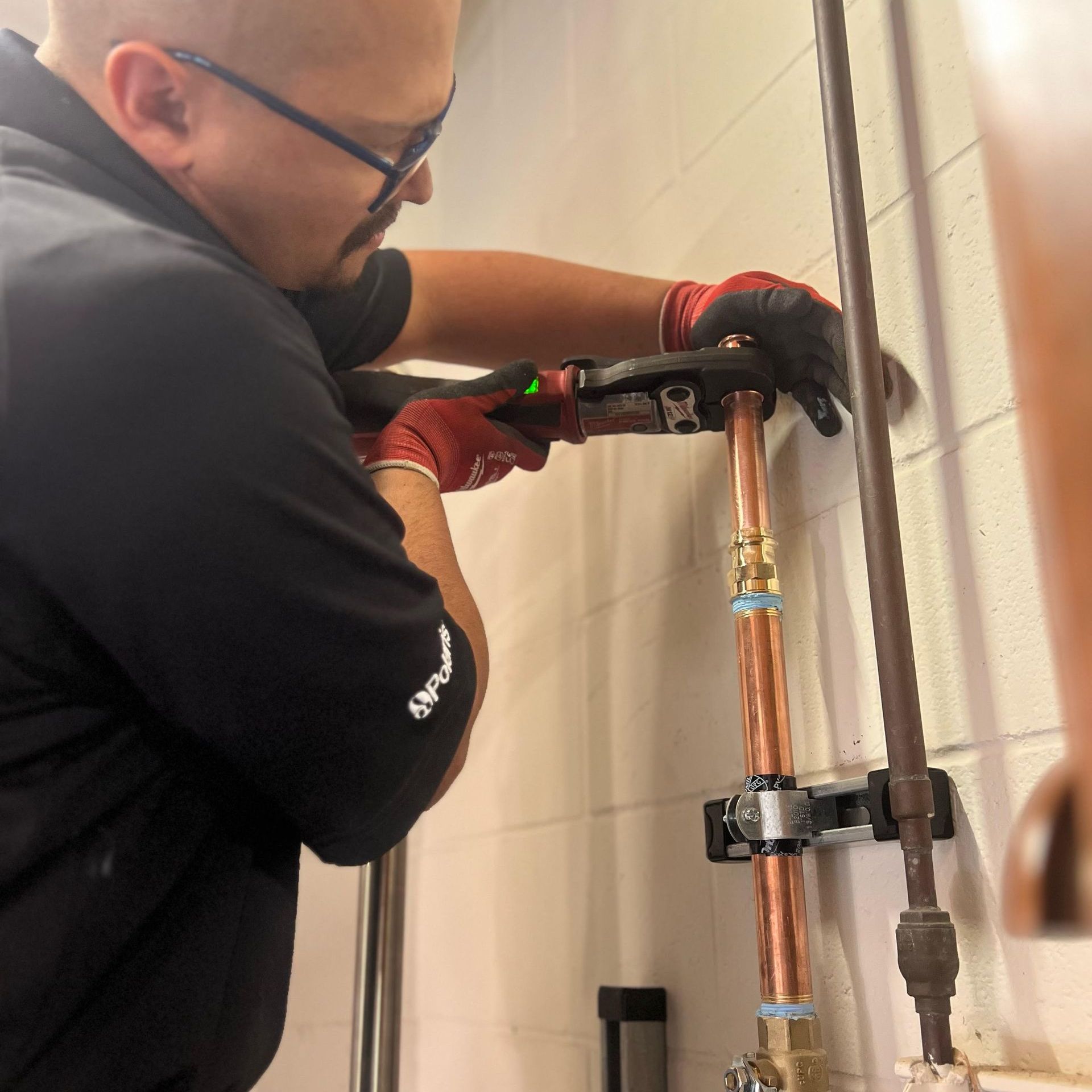 A worker in a black shirt and red gloves uses a power crimping tool to connect copper plumbing pipes on a white wall.