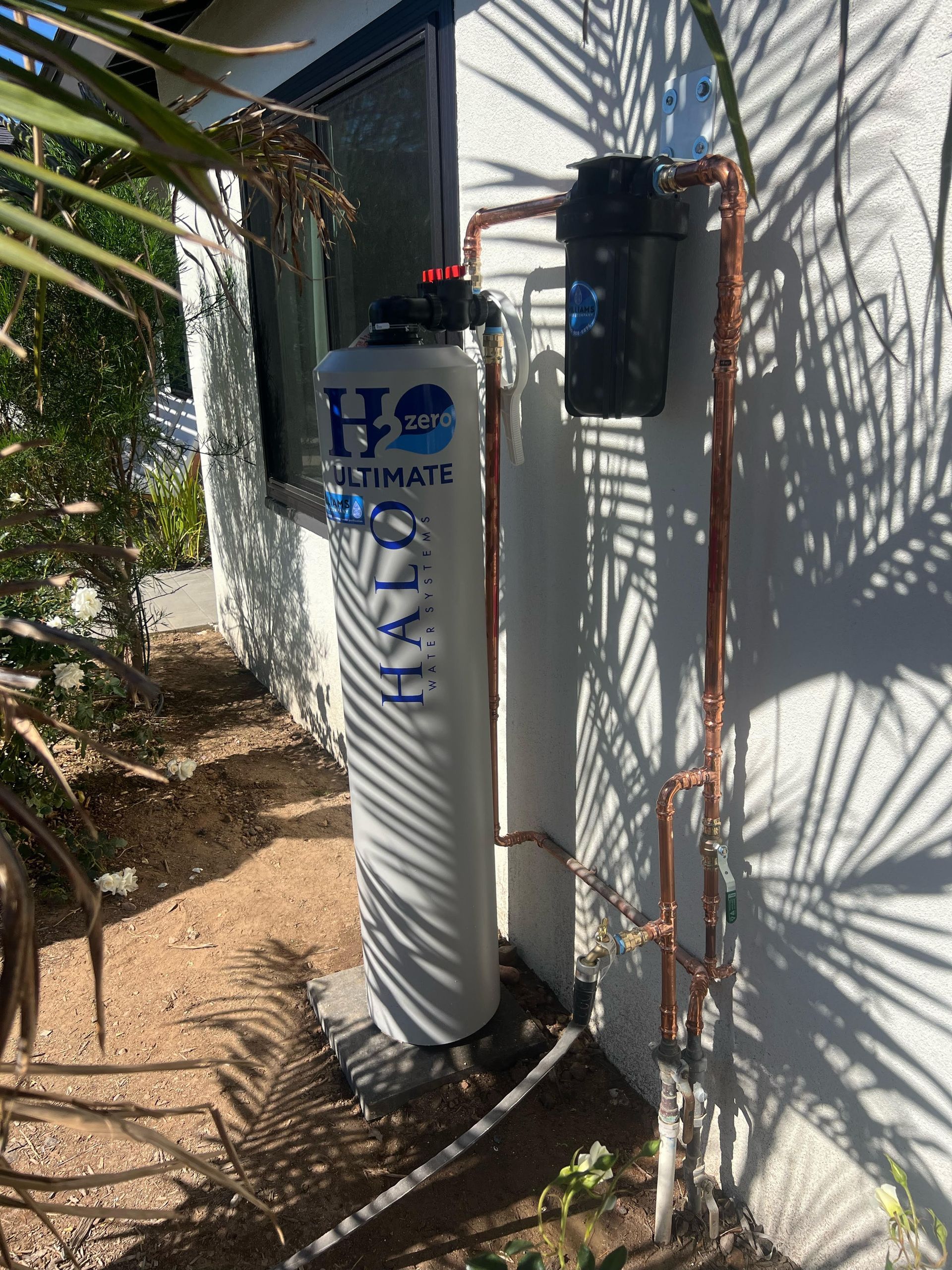 A white Halo water filtration tank and a black pre-filter housing mounted on a white exterior wall next to a window.